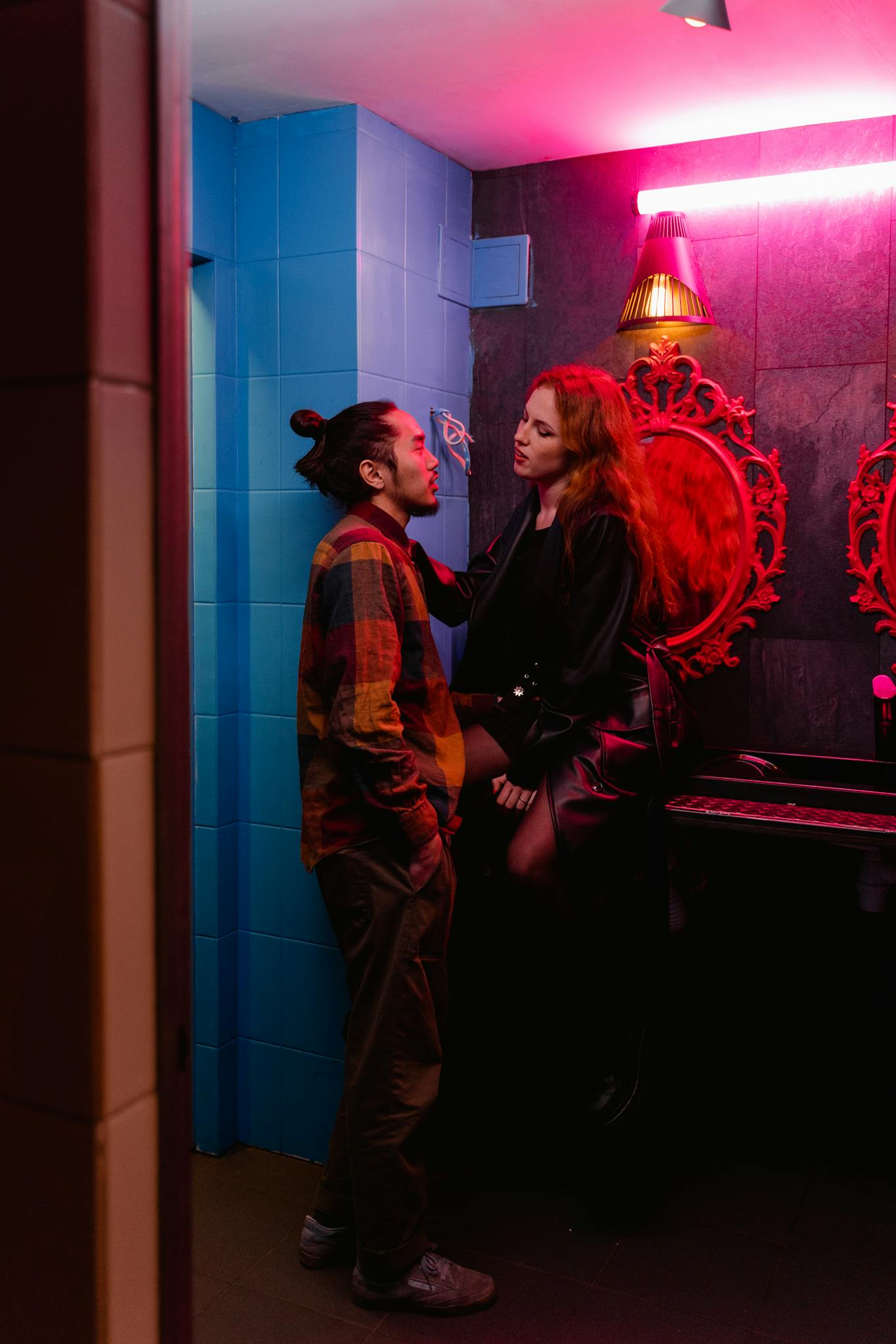 A stylish couple shares a moment in a neon-lit restroom, creating a vibrant nightlife scene.